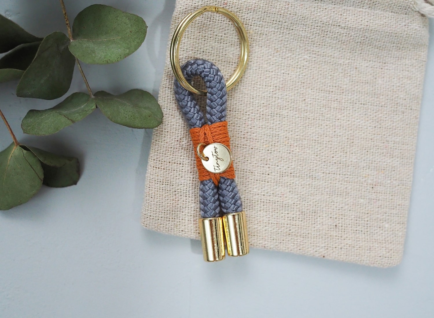 Tau Schlüsselanhänger in grau mit orangefarben Details auf Baumwollsäckchen.