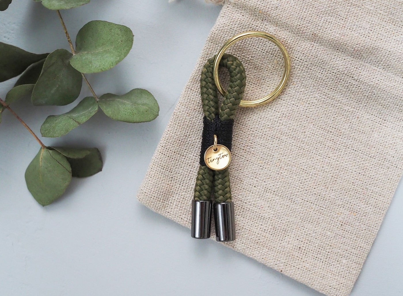 Schlüsselanhänger aus Tau in olivgrün und schwarz mit goldenen Details und beigem Baumwollsäckchen.